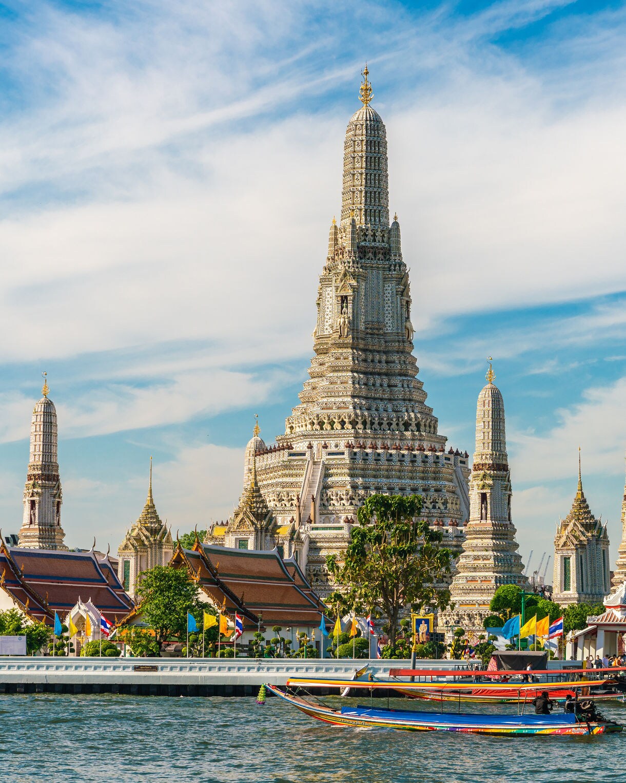 View of Wat Arun temple complex with ornate spires rising behind colorful longtail boats on the Chao Phraya River under a blue sky.