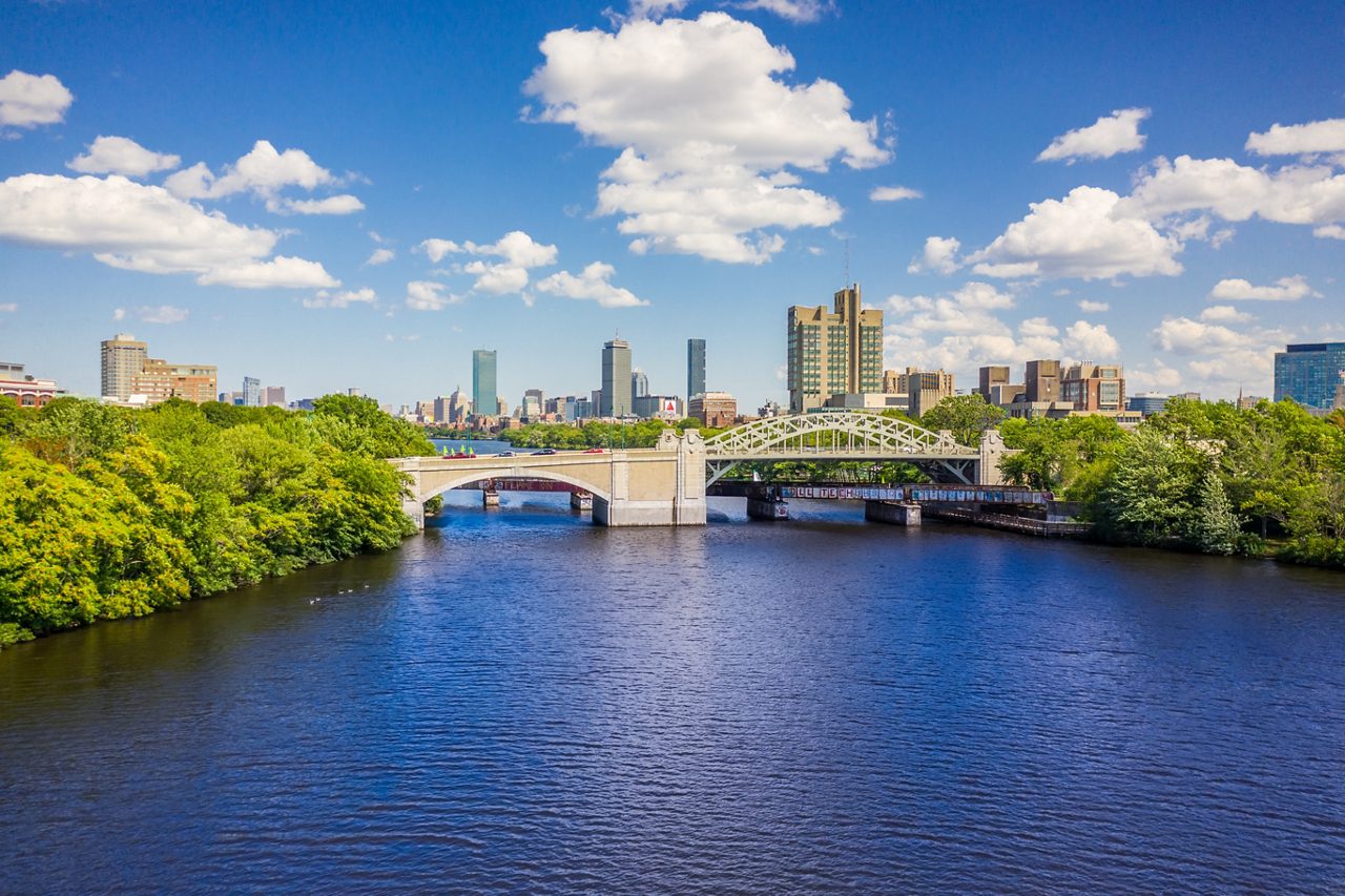 A sunny view of the Charles River with tree-lined banks, twin bridges and Boston’s downtown skyline rising in the distance under a bright blue sky.