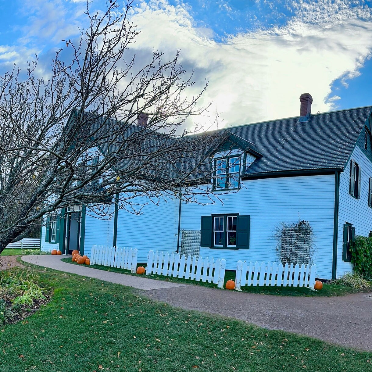 A white farmhouse with green shutters and a dark roof stands behind a white picket fence lined with pumpkins, with a bare tree, green lawn and flower beds under a partly cloudy blue sky.