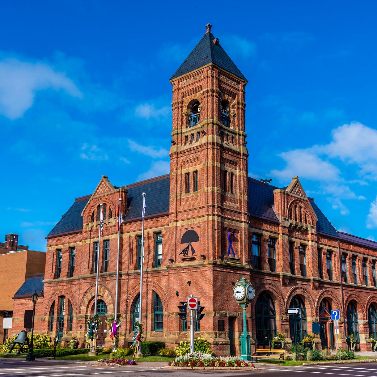 A large red-brick city hall with arched windows, a tall clock tower, flags, and a small landscaped plaza, all set beneath a bright blue sky with scattered clouds.