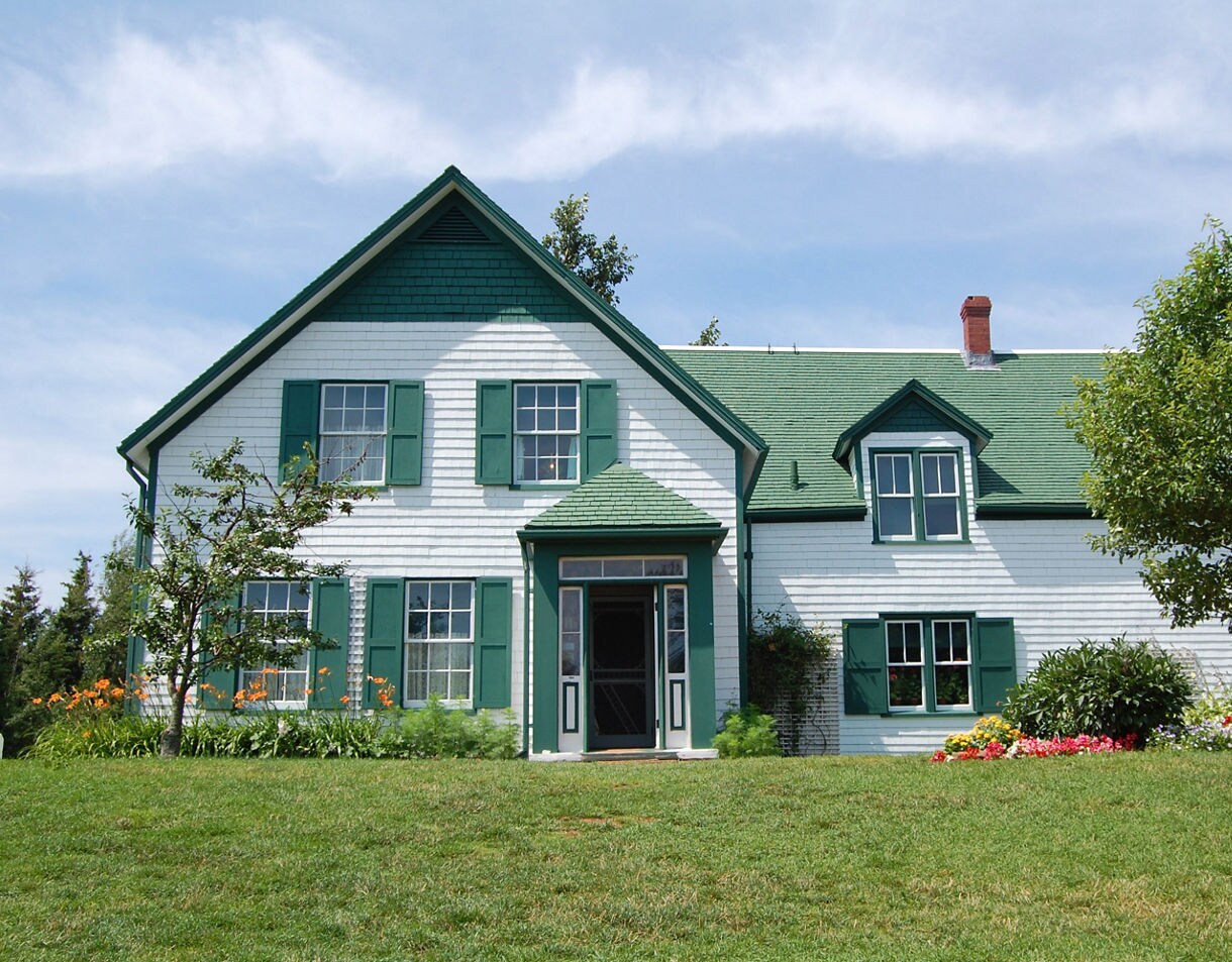 A white farmhouse with green shutters and a green roof, set on a grassy lawn with colorful flower beds and a few trees under a clear blue sky.