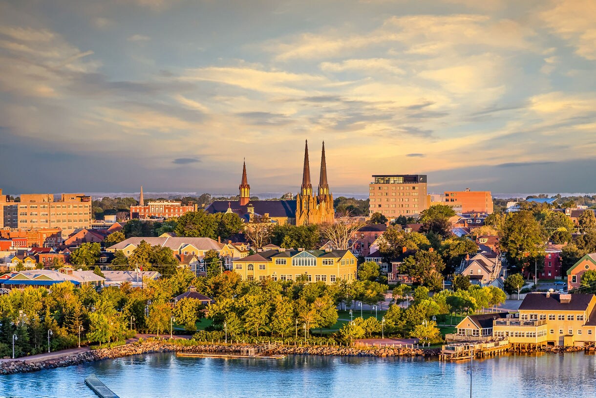 A panoramic view of Charlottetown at sunset, showing tall church spires, colorful homes, mid-rise buildings and a tree-filled waterfront beside calm harbor water under a warm sky.