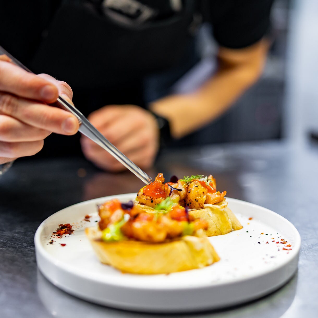 A close-up of a chef using metal tweezers to place garnishes on small seafood-topped bruschetta pieces arranged on a white plate in a professional kitchen.