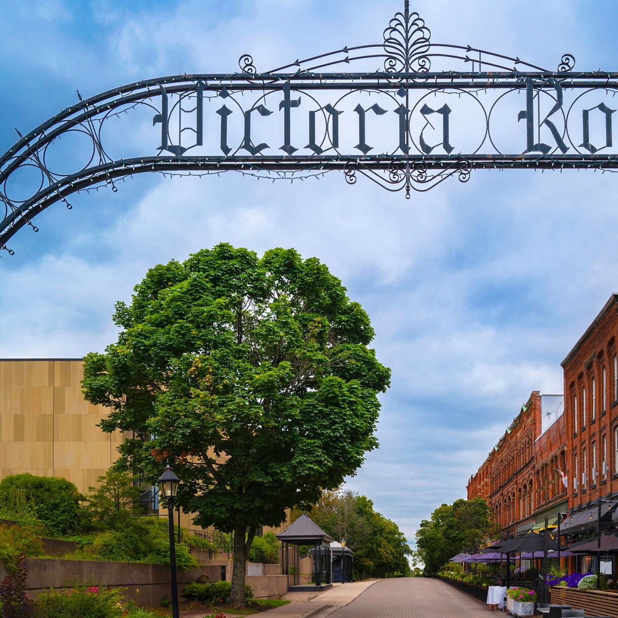 An ornate black metal arch reading “Victoria Row” spans above a pedestrian street lined with tall brick buildings, outdoor restaurant patios, and green trees under a cloudy sky.