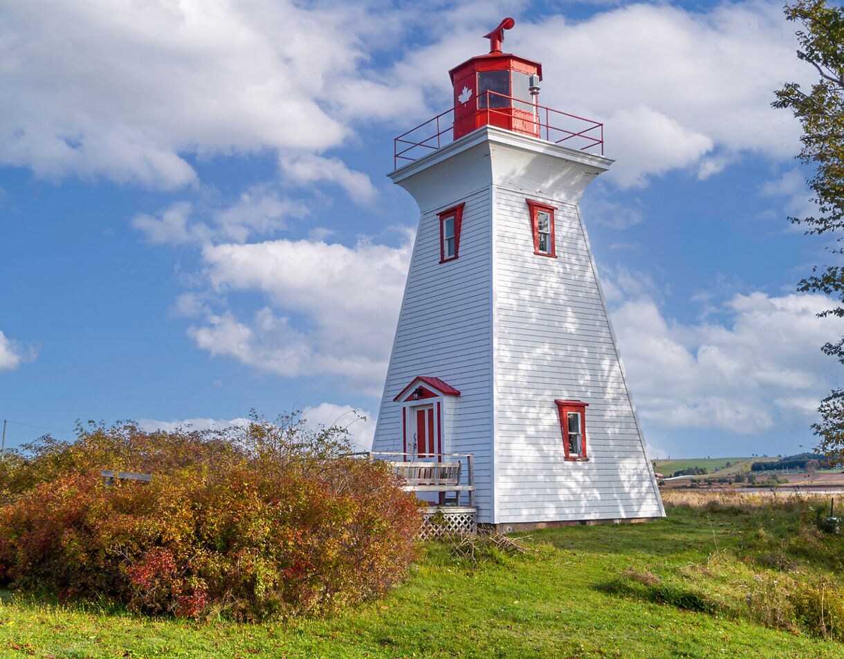 A white wooden lighthouse with red trim and a red lantern room, set on a grassy field with autumn shrubs under a blue sky filled with scattered clouds.
