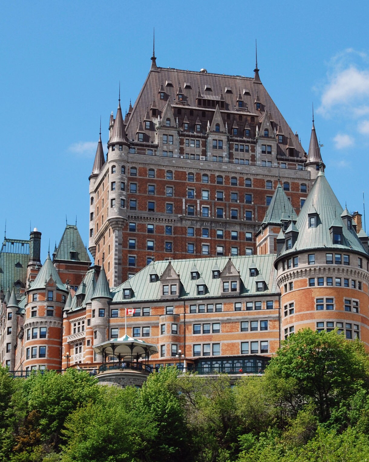 Château Frontenac rising over green trees, showing its copper roofs, brick façade and tall central tower beneath a bright blue sky.