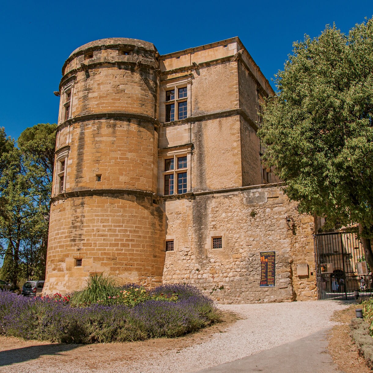 Stone tower and walls of Château de Lourmarin surrounded by trees, flowers and a bright summer sky in southern France.