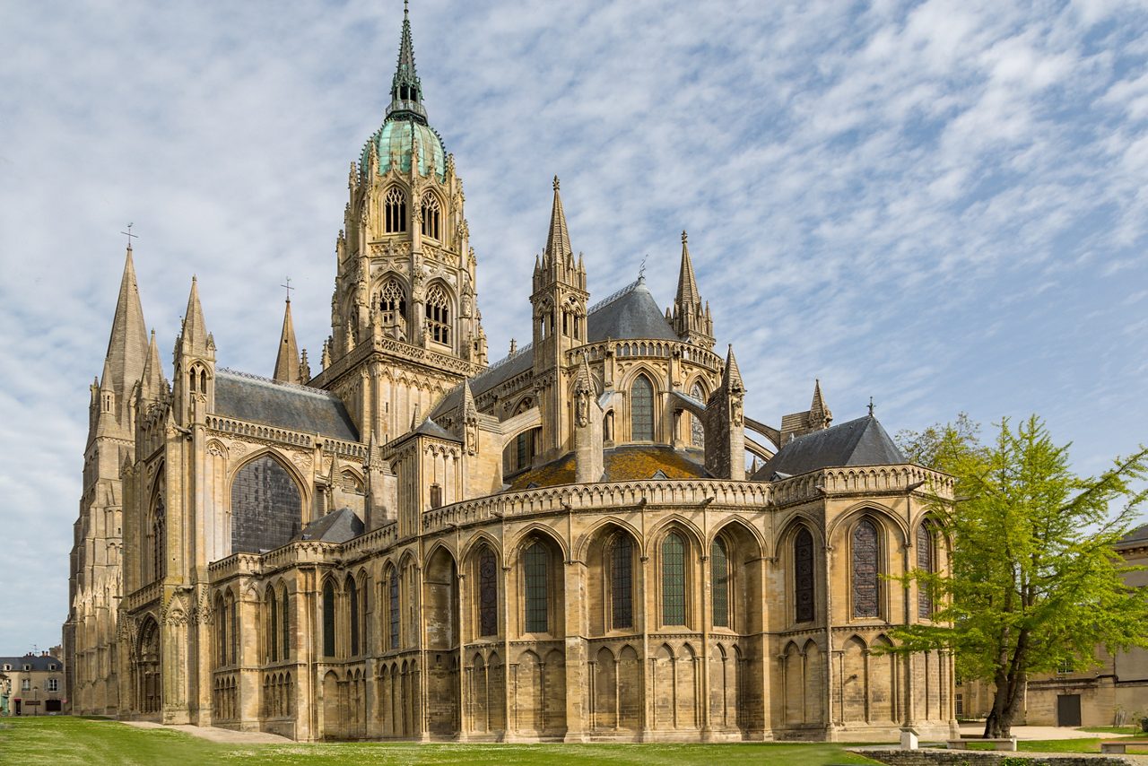 Exterior view of Bayeux Cathedral in Normandy with tall stone towers, arched windows and intricate detailing under a partly cloudy sky.