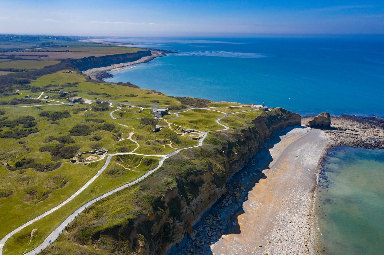 Aerial view of Pointe du Hoc in Normandy showing grassy bunkers, winding paths and steep cliffs dropping into the English Channel.