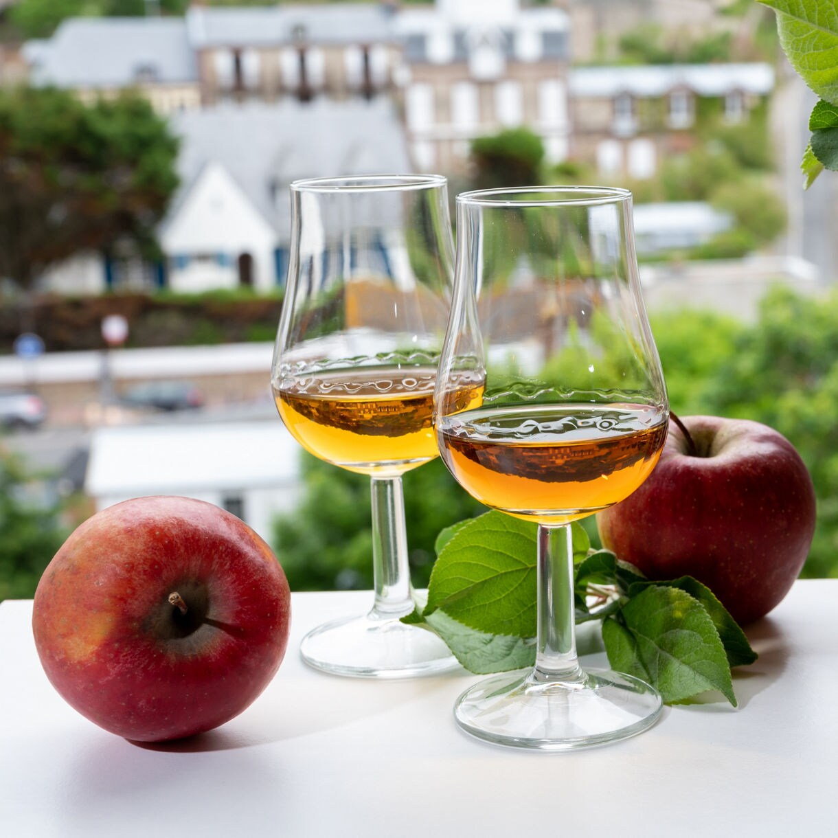 Two glasses of amber apple brandy beside fresh red apples and a wicker basket, set against a blurred village backdrop.