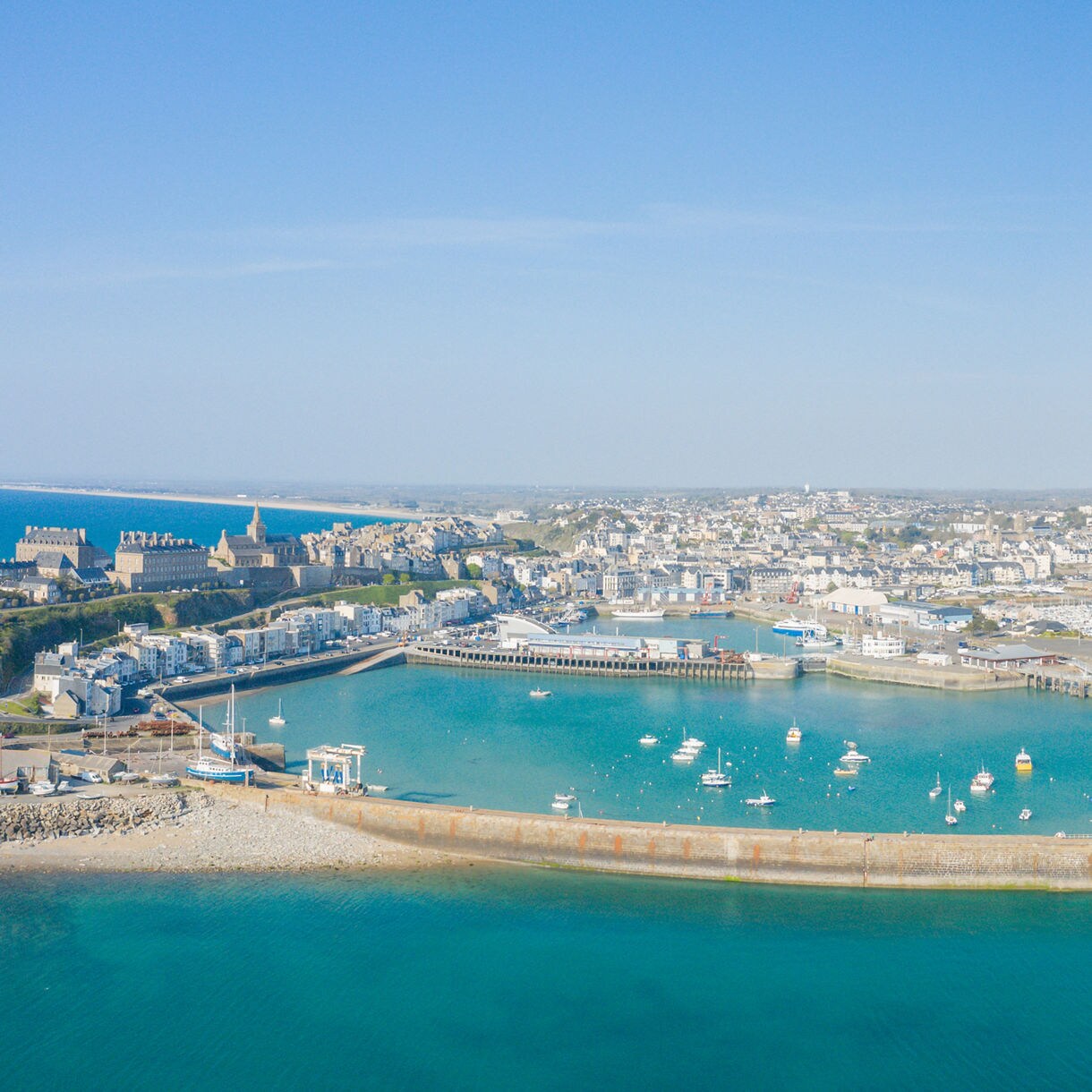  Aerial view of Cherbourg harbor with a curved sea wall, moored boats and the town’s historic buildings stretching along the coastline under a clear blue sky.