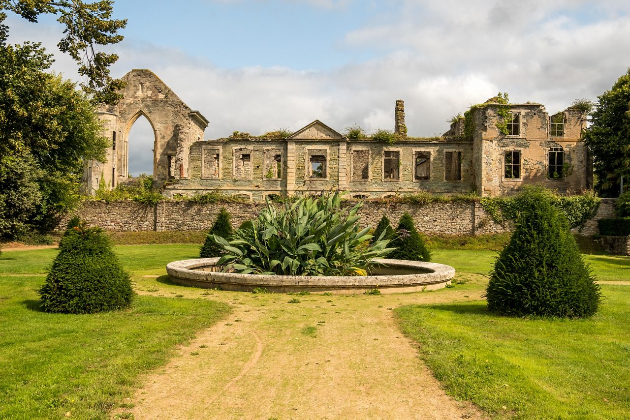 Weathered stone ruins with empty window frames and a tall arched opening, framed by manicured lawns and a circular garden planter in the foreground.