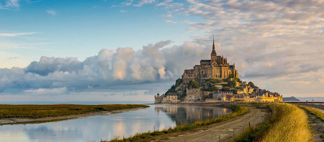 Mont Saint-Michel abbey perched on a rocky island, surrounded by reflective tidal flats and dramatic clouds at sunset.