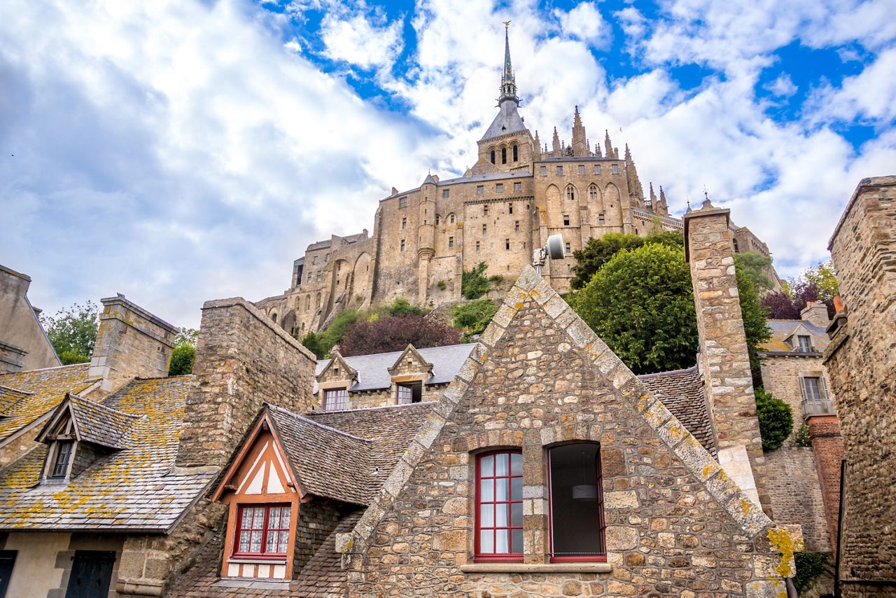 View from below of Mont Saint-Michel’s stone abbey rising over rustic village rooftops with timber and stone details against a partly cloudy sky.
