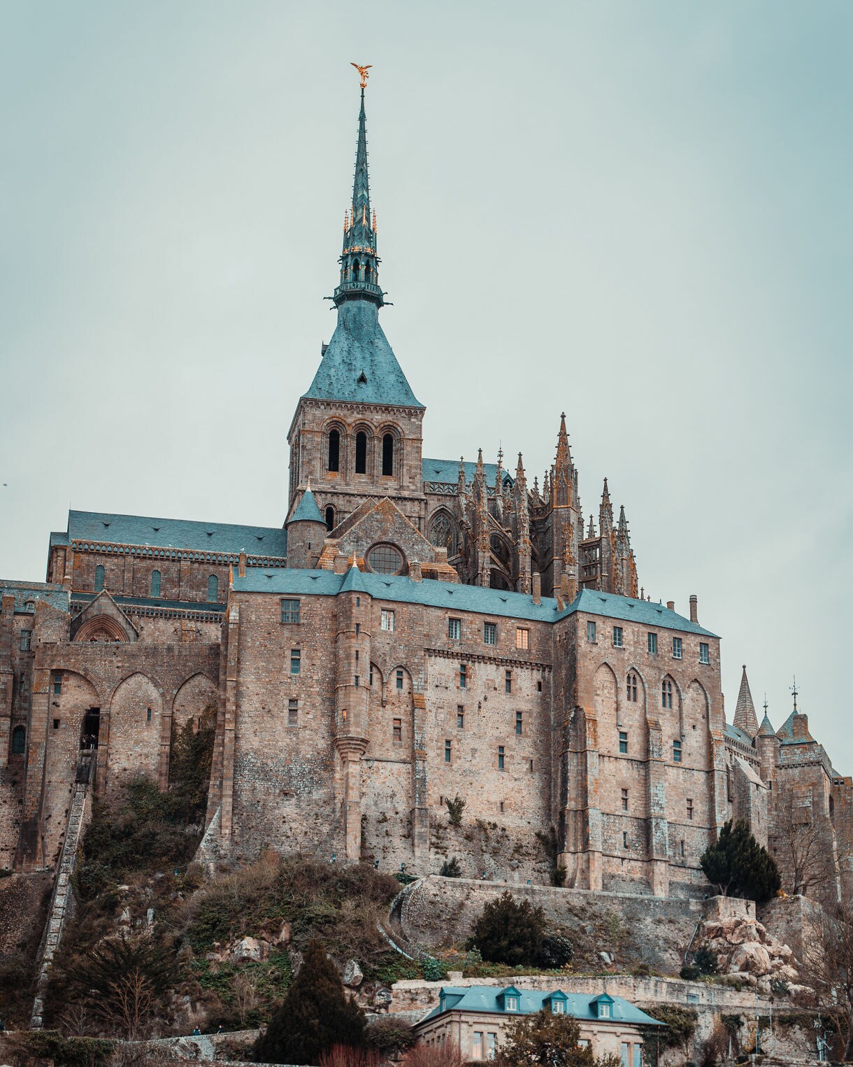 Vertical view of Mont Saint-Michel’s stone abbey rising above village rooftops with a tall spire topped by a golden statue against a cloudy sky.