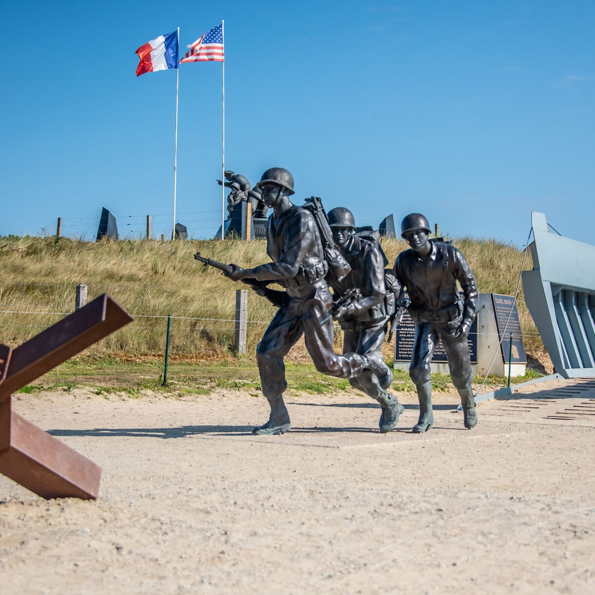 Bronze statues of WWII soldiers charging onto Normandy beach near a landing craft replica, with French and American flags waving overhead.