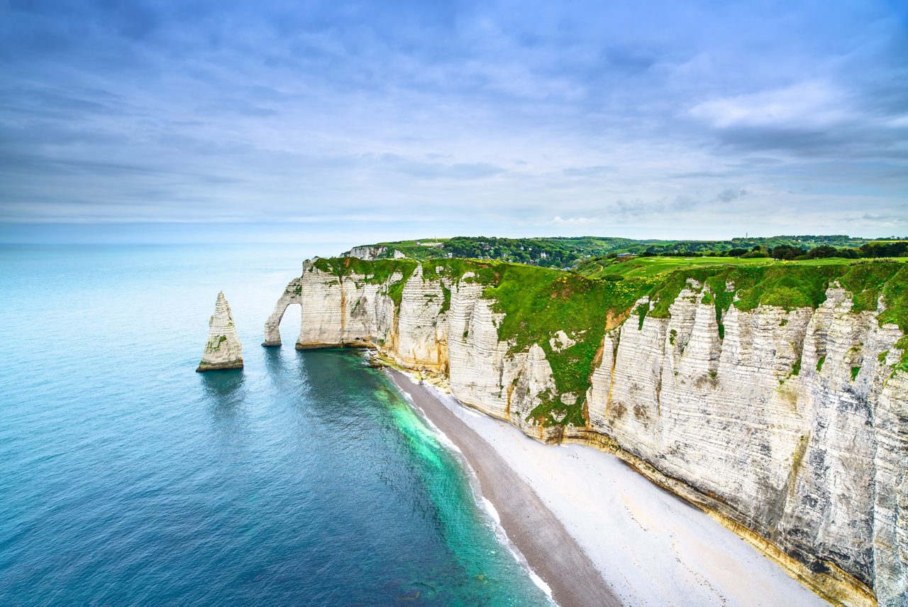 Dramatic white chalk cliffs of Étretat in Normandy with a natural arch and pointed sea stack rising above turquoise waters under a cloudy sky.