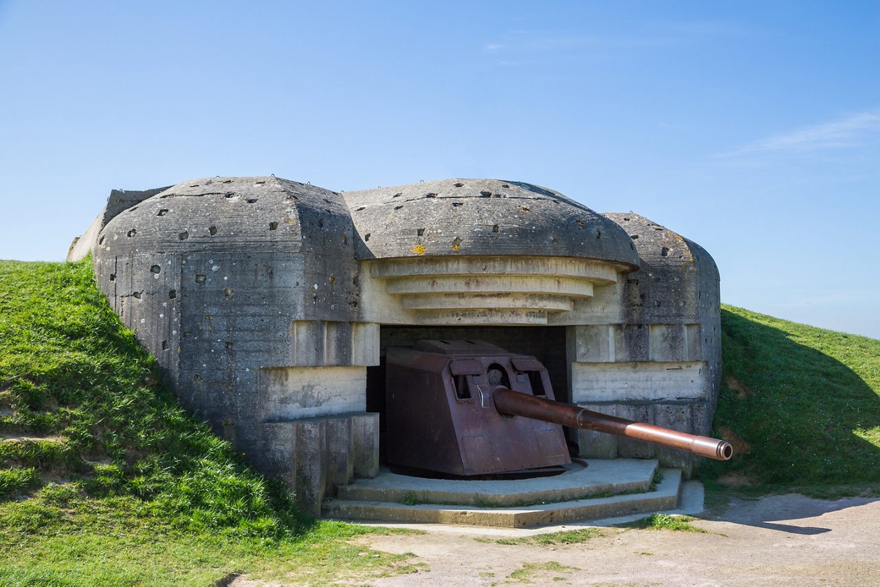  Concrete German bunker from World War II housing a preserved naval gun, set into grassy earthworks under a clear blue sky.