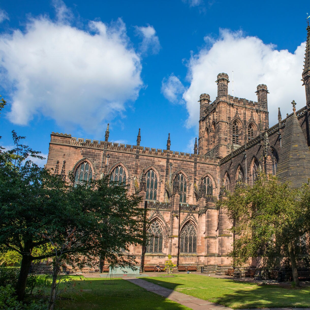 Chester Cathedral in England with tall Gothic spires, arched windows and stone walls framed by trees and greenery.