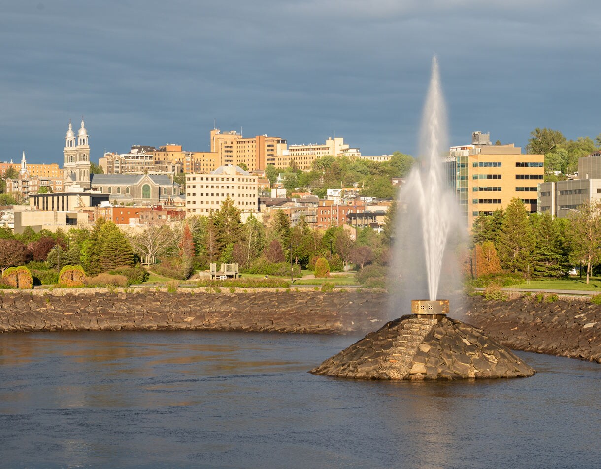 Riverside view of Chicoutimi with a tall fountain in the foreground and city buildings and trees in the background under soft golden light.