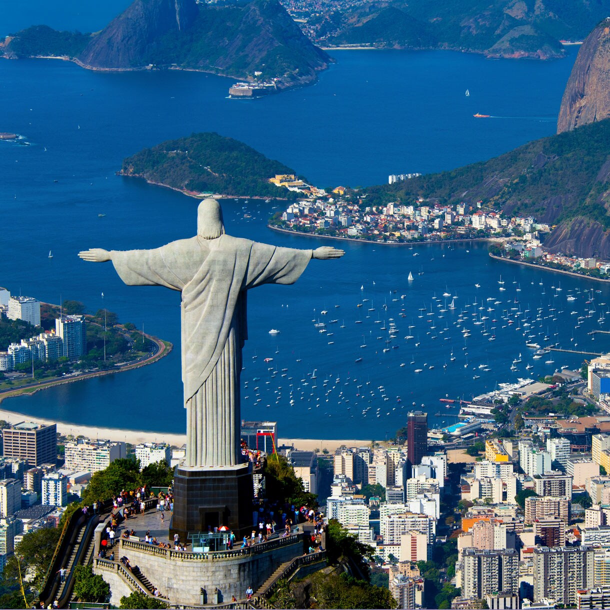 Aerial scene of Christ the Redeemer overlooking Rio de Janeiro, with Guanabara Bay full of boats and Sugarloaf Mountain rising prominently in the background.