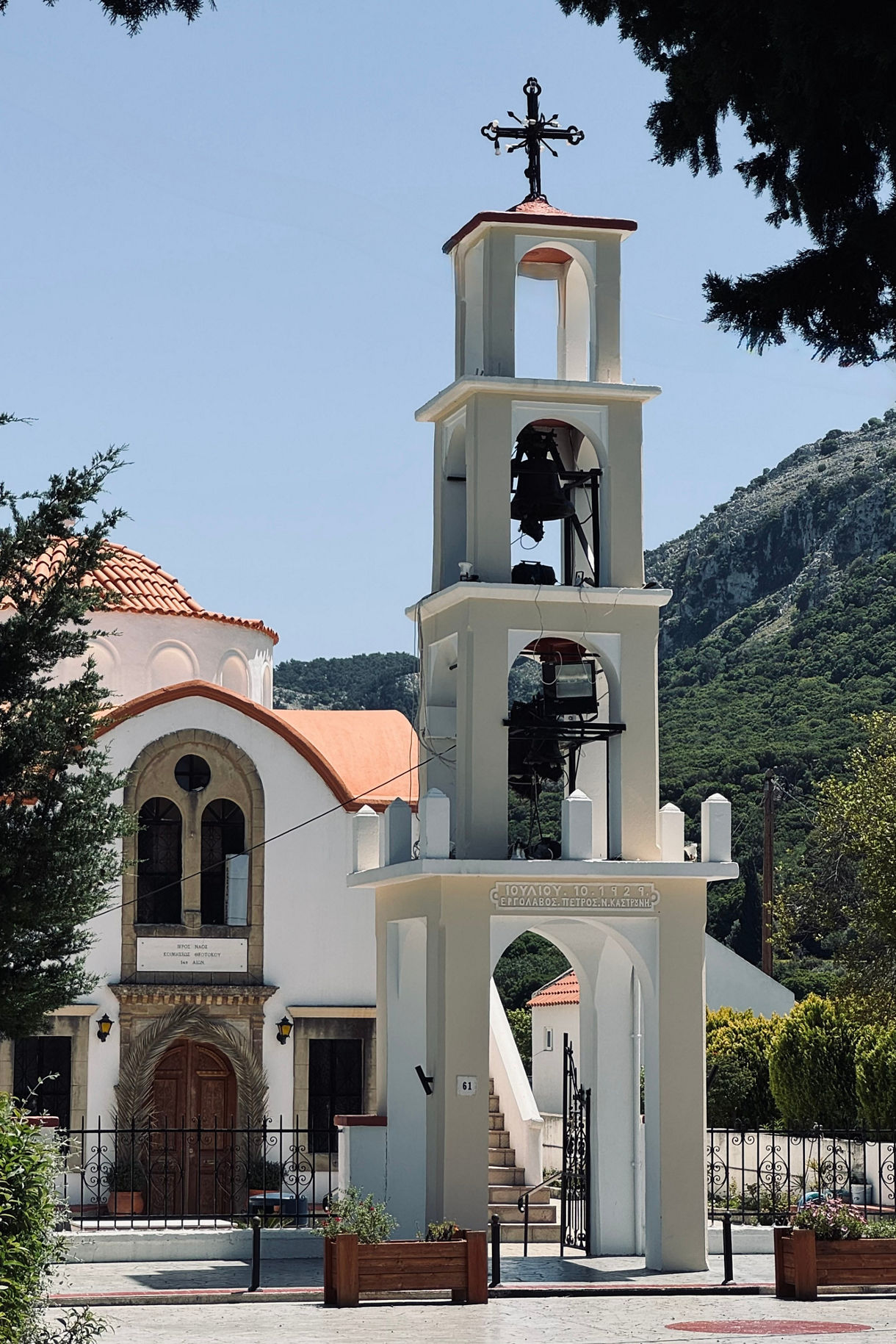 Whitewashed Church of the Assumption with a red-tiled dome and bell tower, set against green hills on a sunny day in Rhodes, Greece.