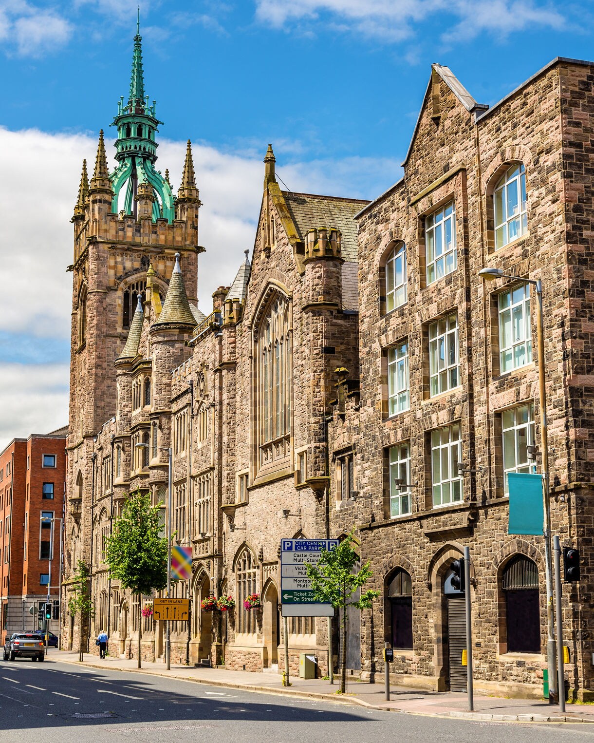Gothic Revival stone building in Belfast known as Church House, featuring arched windows, detailed stonework and a tall green spire rising above the street.