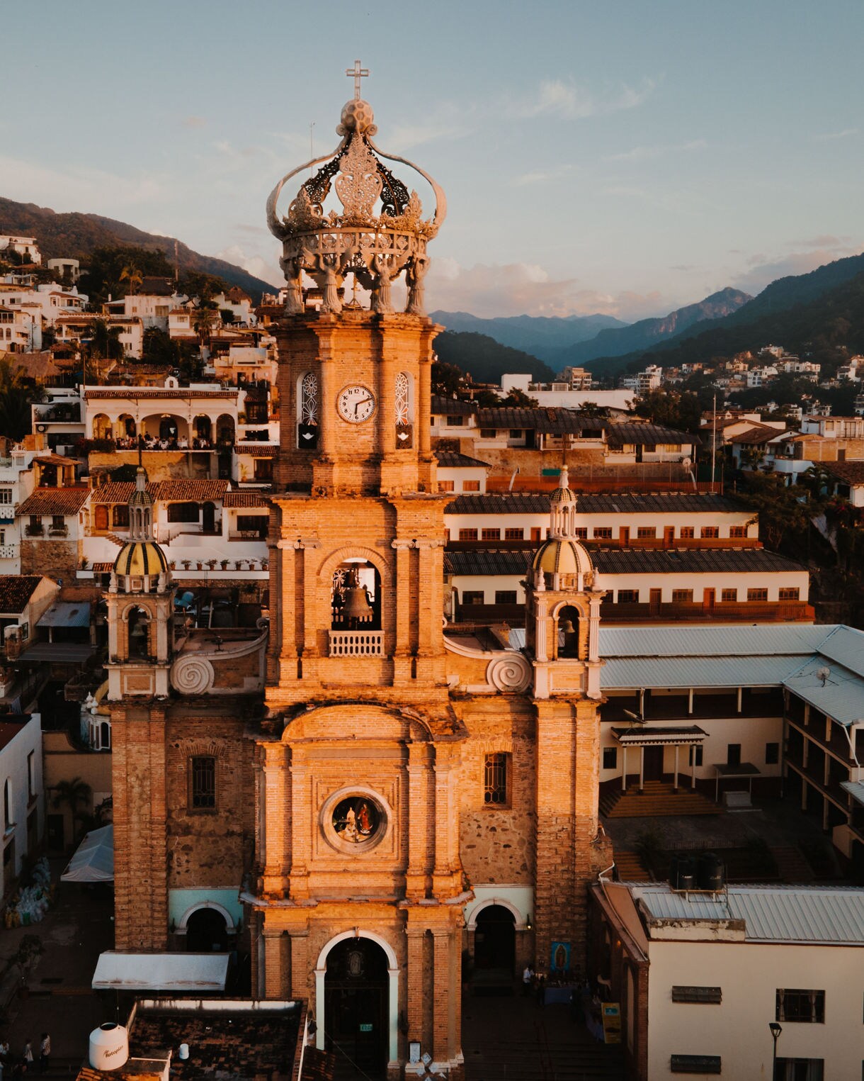 Sunset view of the Church of Our Lady of Guadalupe in Puerto Vallarta, Mexico, with its ornate crown-topped bell tower surrounded by hillside houses and mountains.