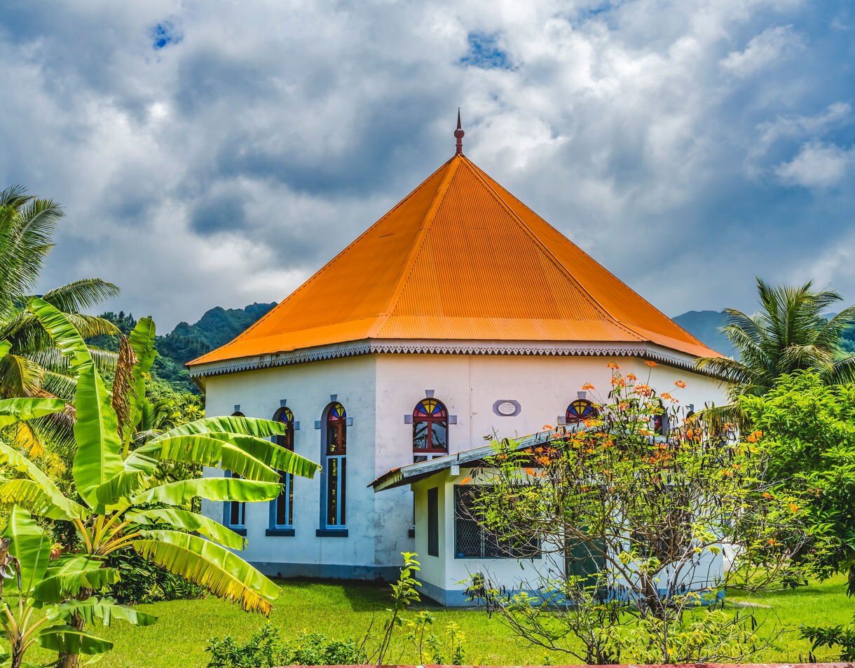 White octagonal church with a bright orange roof in Papetoai, Moorea, surrounded by palm trees and lush tropical greenery.