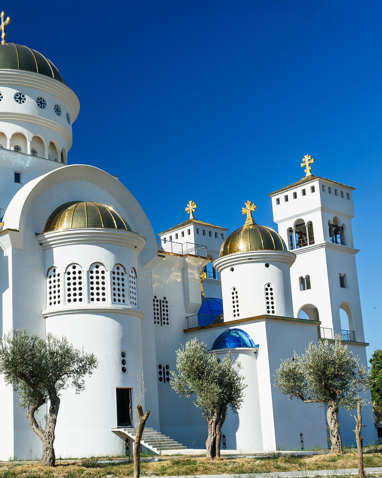White Orthodox church in Bar, Montenegro with multiple gold domes and crosses, viewed under a deep blue sky with olive trees in the foreground.
