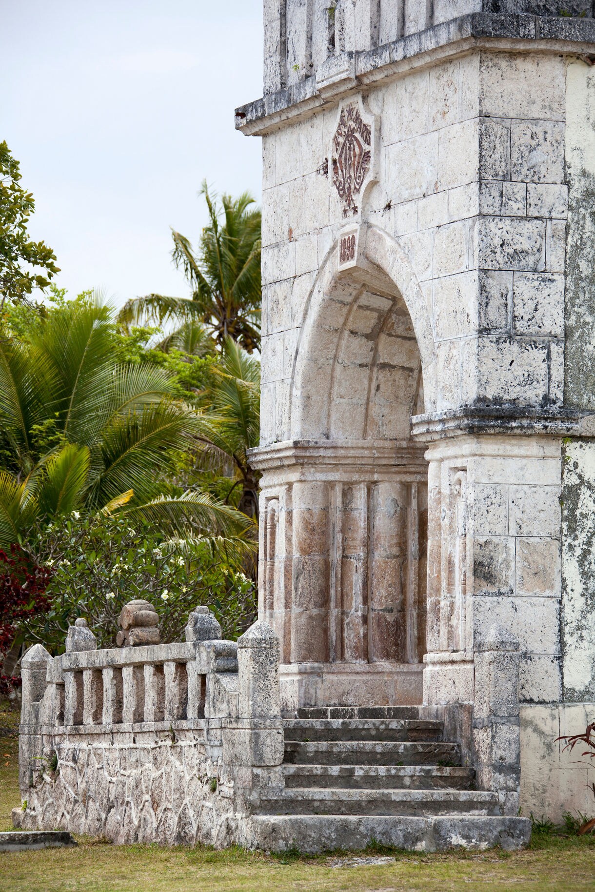 Close view of an old stone church entrance with carved arches and worn steps, surrounded by tropical greenery and palm trees.