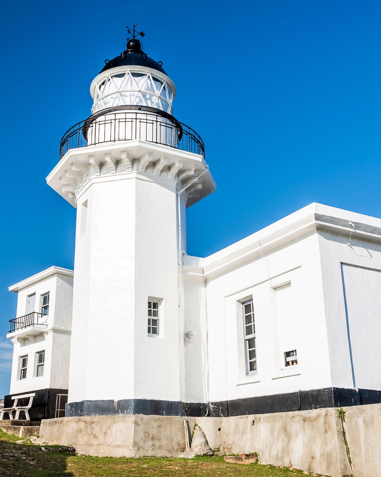  White Cihou Lighthouse in Kaohsiung, Taiwan, standing under a clear blue sky with two flags flying nearby and sunlight highlighting its structure.