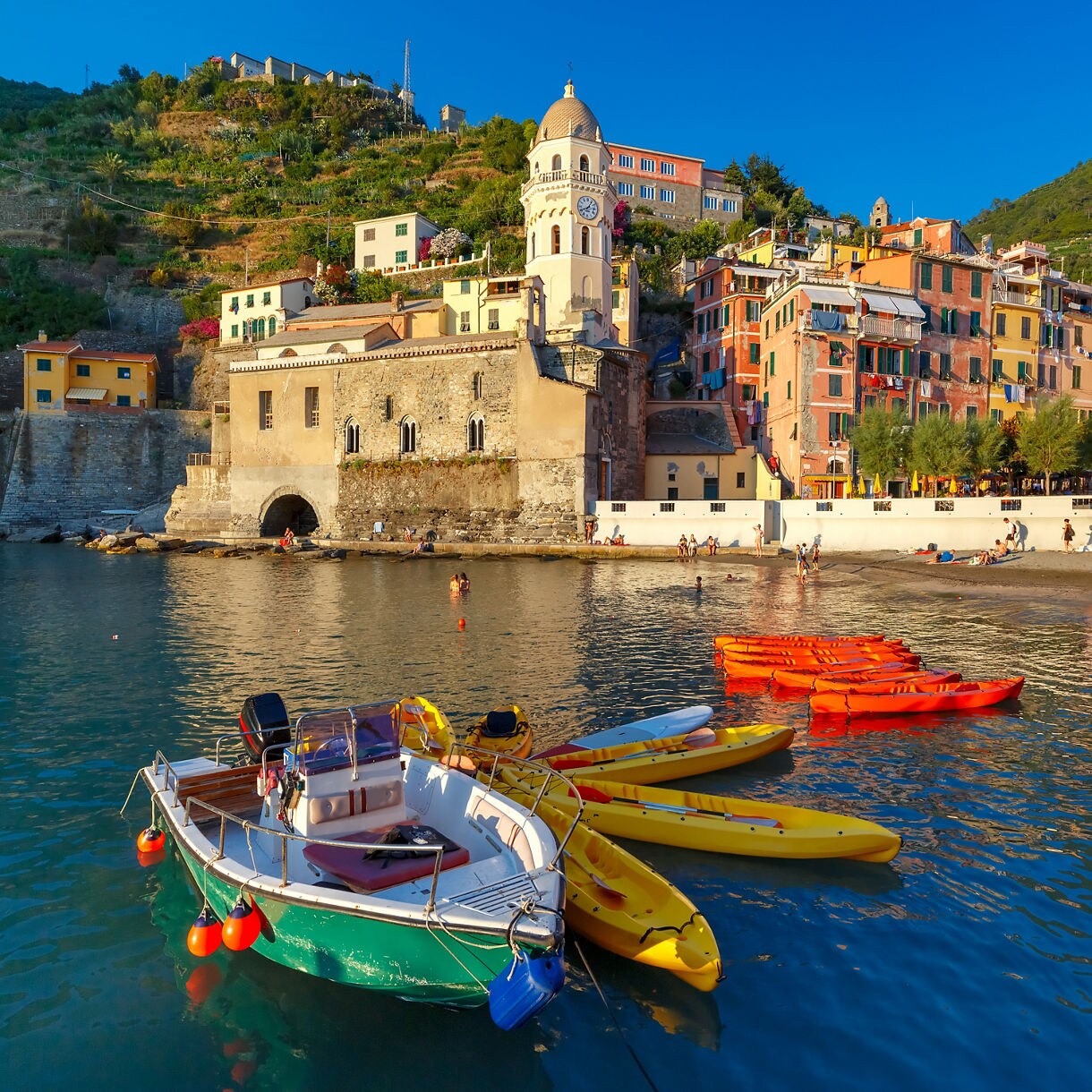 A small harbor in Vernazza with colorful kayaks and a boat floating in calm water, backed by pastel hillside buildings and a church by the shoreline.