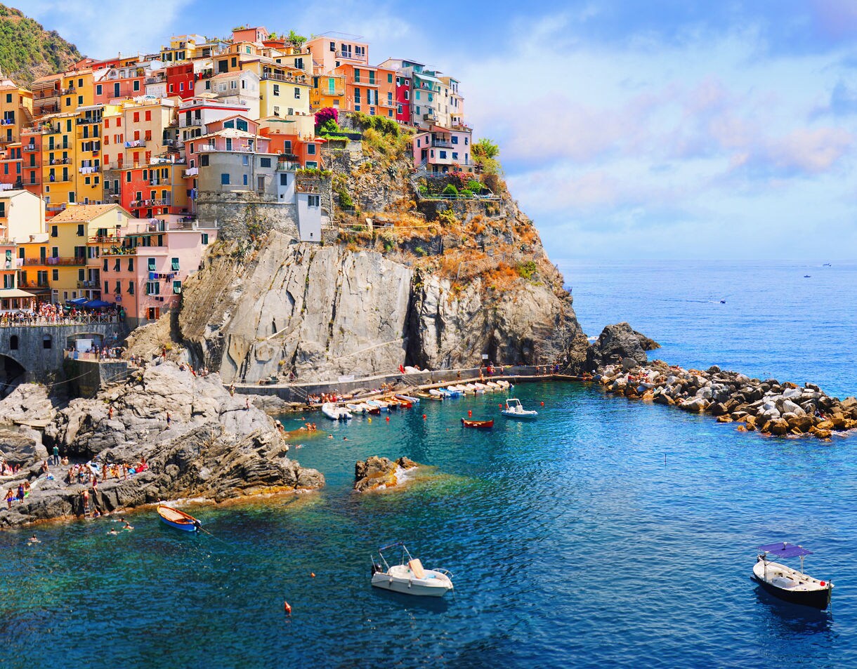 Panoramic view of Manarola in Cinque Terre, Italy, showing vibrant hillside houses above rocky cliffs and turquoise water dotted with small boats.
