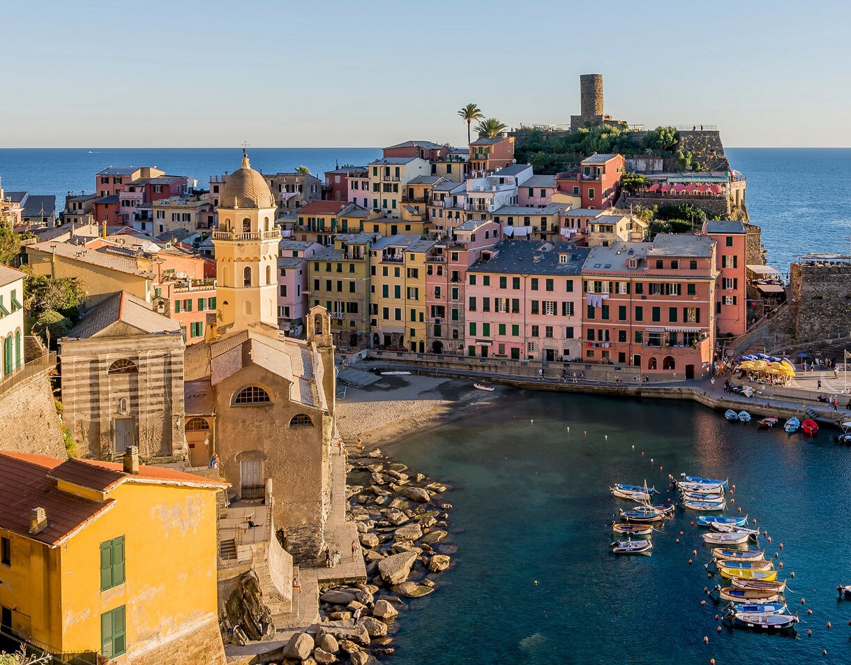 A coastal village with pastel houses stacked along a hillside, a church with a domed bell tower near the waterfront and small fishing boats floating in a sheltered turquoise harbor.