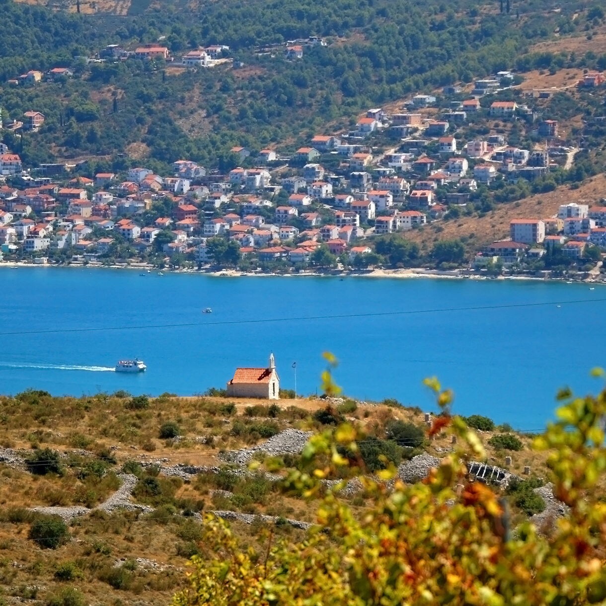 Small stone chapel with a red roof on a dry hillside overlooking bright blue water, with a coastal town of white houses and red rooftops across the bay.