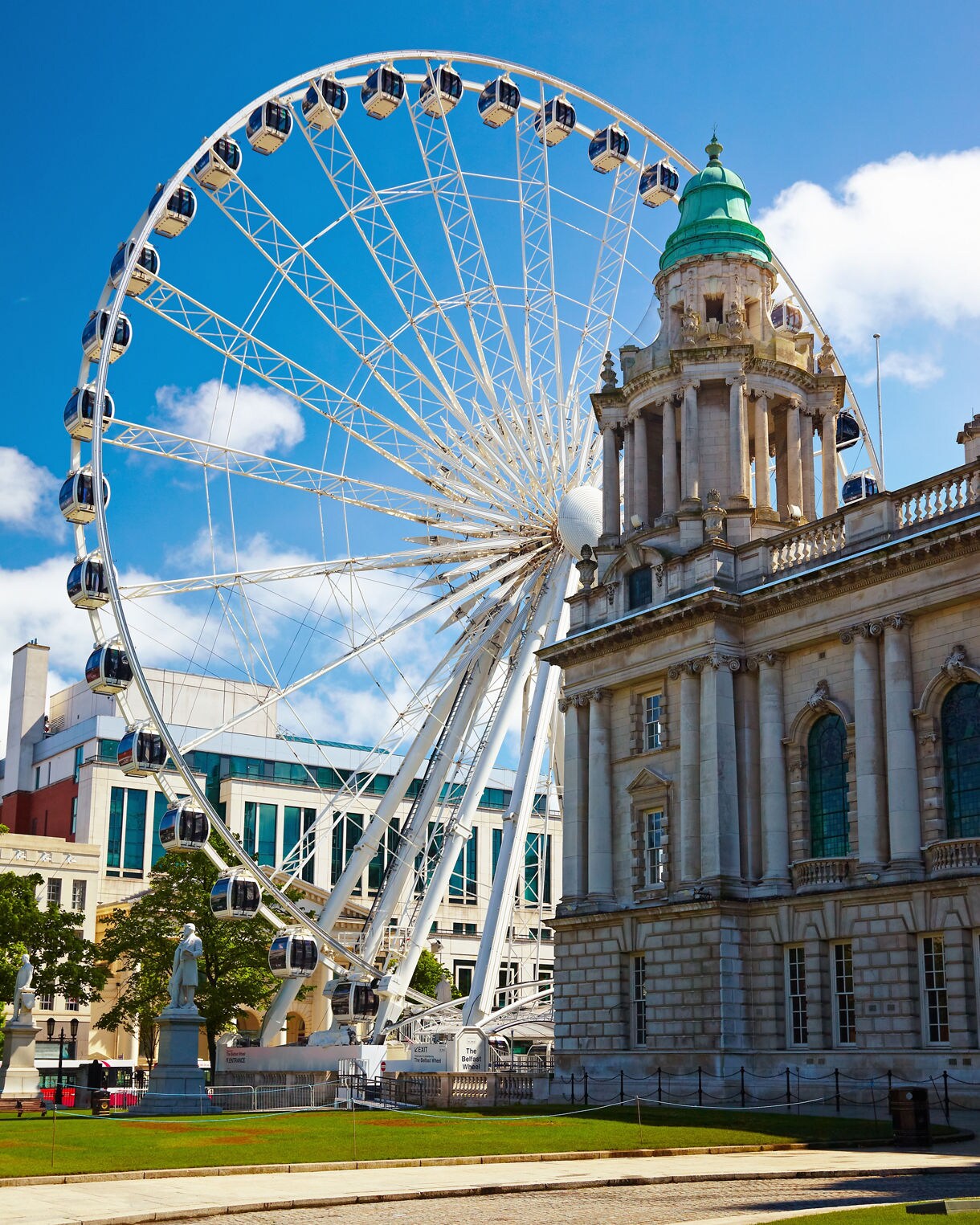 Large Ferris wheel next to Belfast City Hall, with the historic stone building’s green dome and statues contrasting against the bright blue sky.
