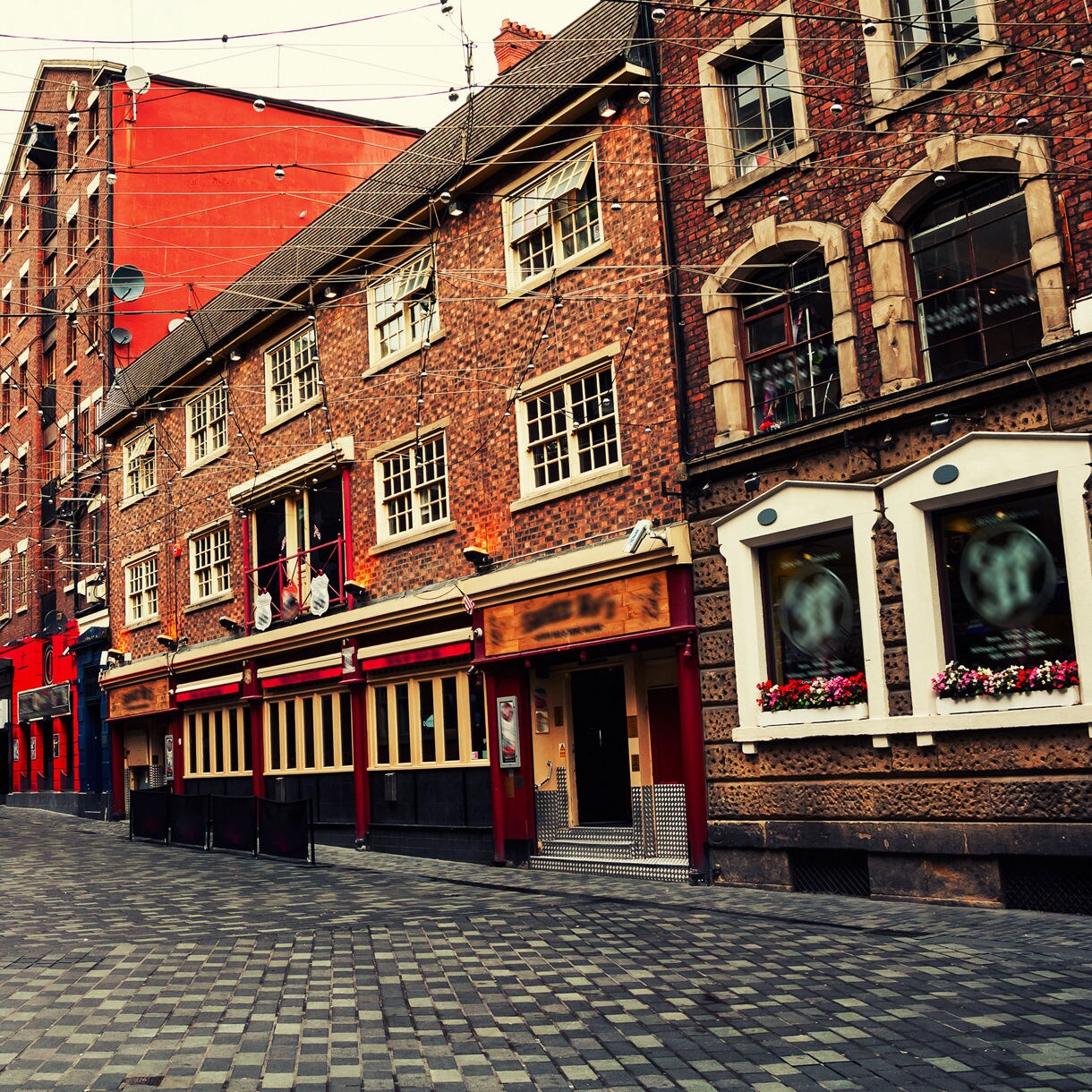 Narrow cobbled street in Liverpool’s city center lined with red-brick buildings, pubs and hanging string lights overhead.