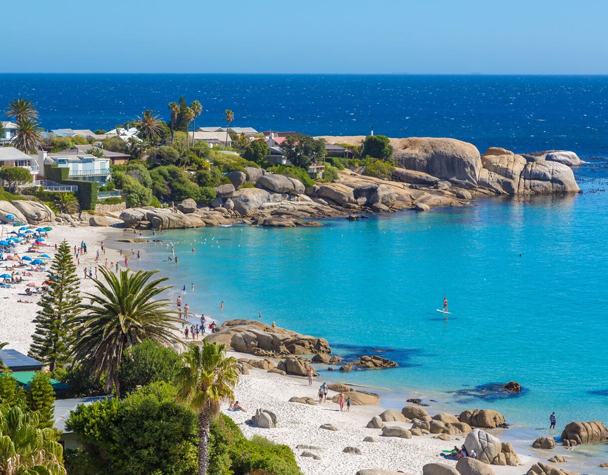 Crowded beach with white sand, turquoise waves, large boulders and houses surrounded by palm trees.