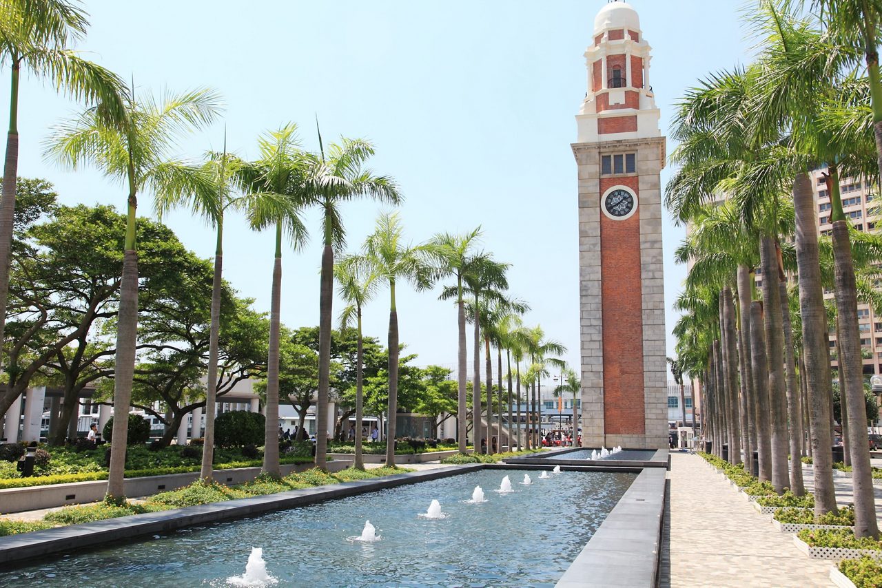 The red-brick Clock Tower in Hong Kong with rows of palm trees and fountains along a reflecting pool under a clear blue sky.