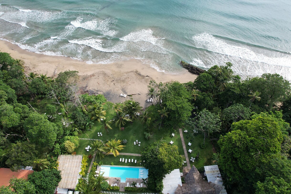 Aerial view of a coastal resort in Limón, Costa Rica with a pool, lush greenery and sandy shoreline meeting ocean waves.