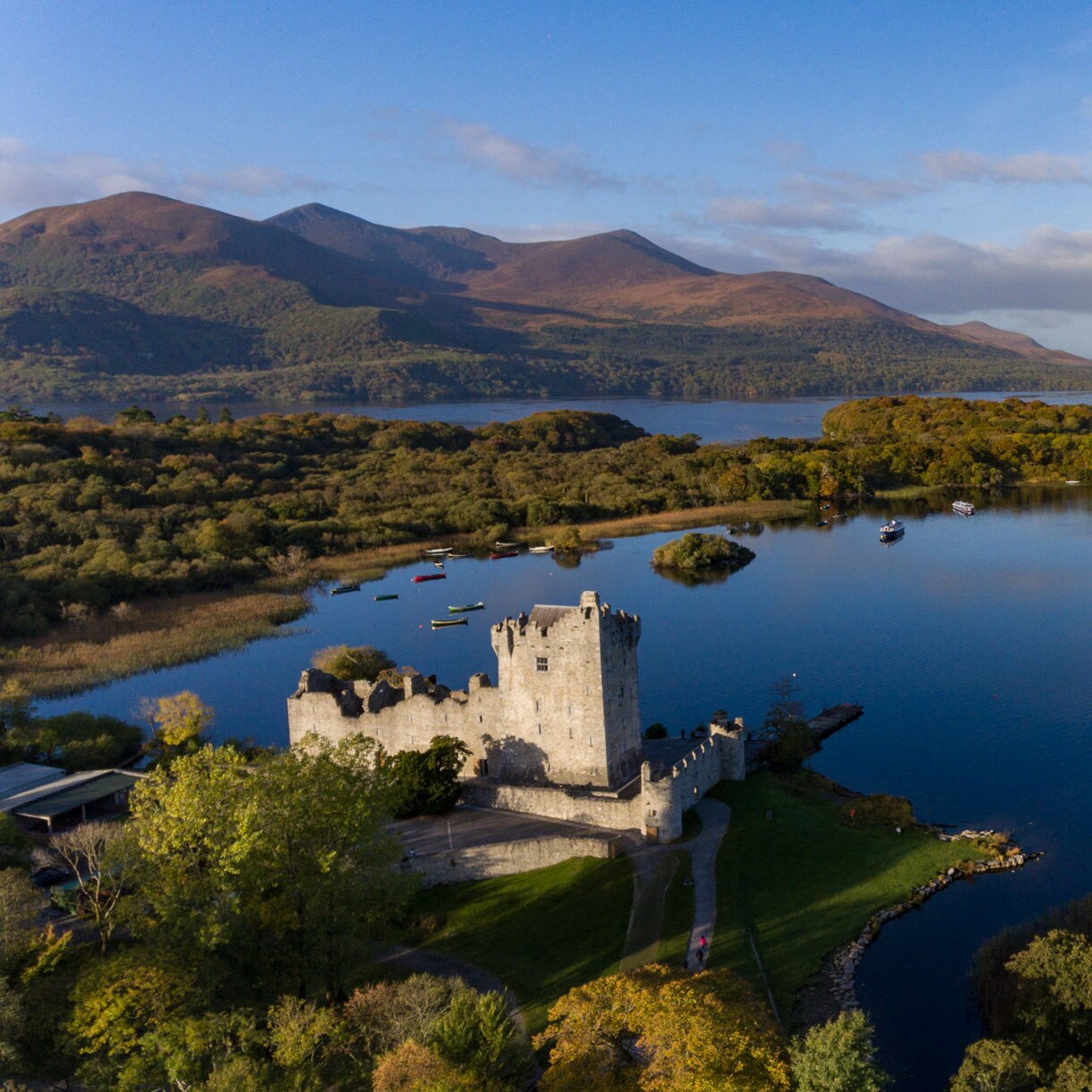 Aerial view of a stone castle beside a wide blue lake with wooded islands and rolling mountains in Killarney National Park.