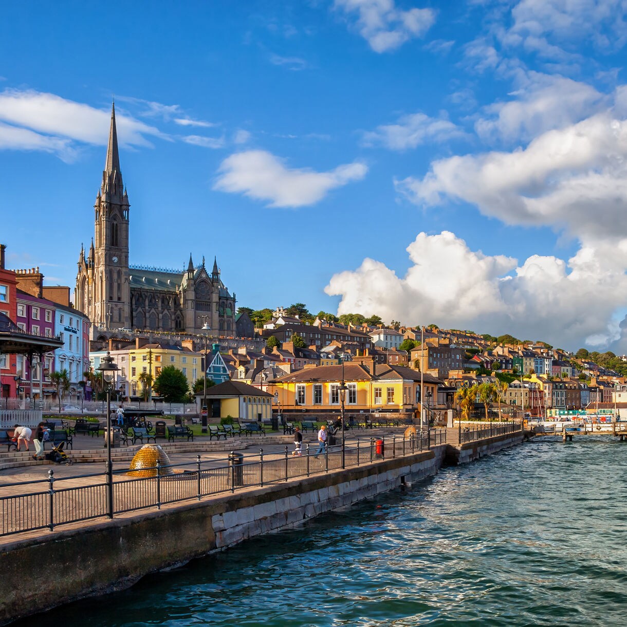 Colorful houses line the waterfront promenade in Cobh with St Colman’s Cathedral rising above the harbor under a bright blue sky with scattered clouds.
