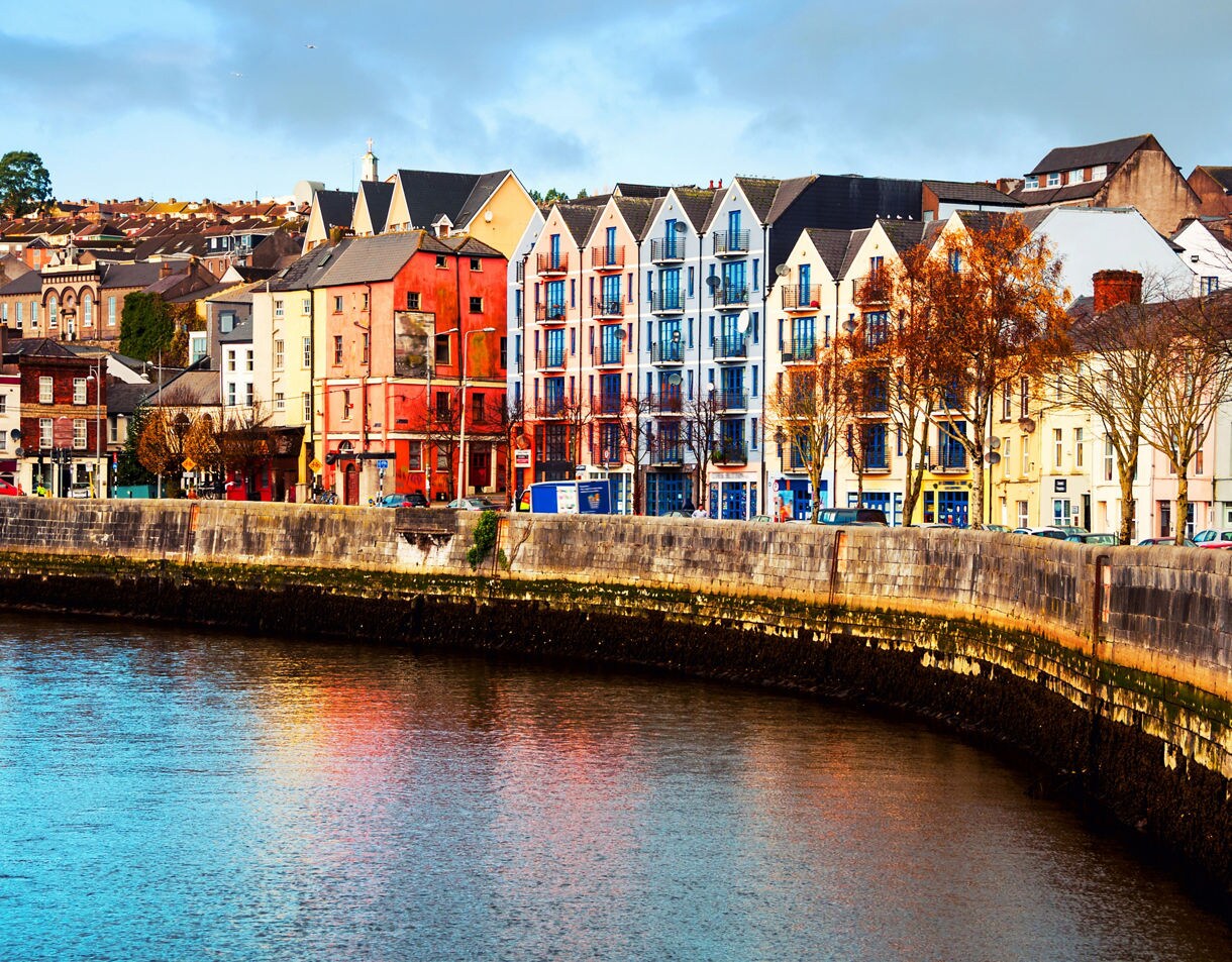 Brightly painted row houses line a curved stone quay along a calm river with reflections shimmering under a blue sky.