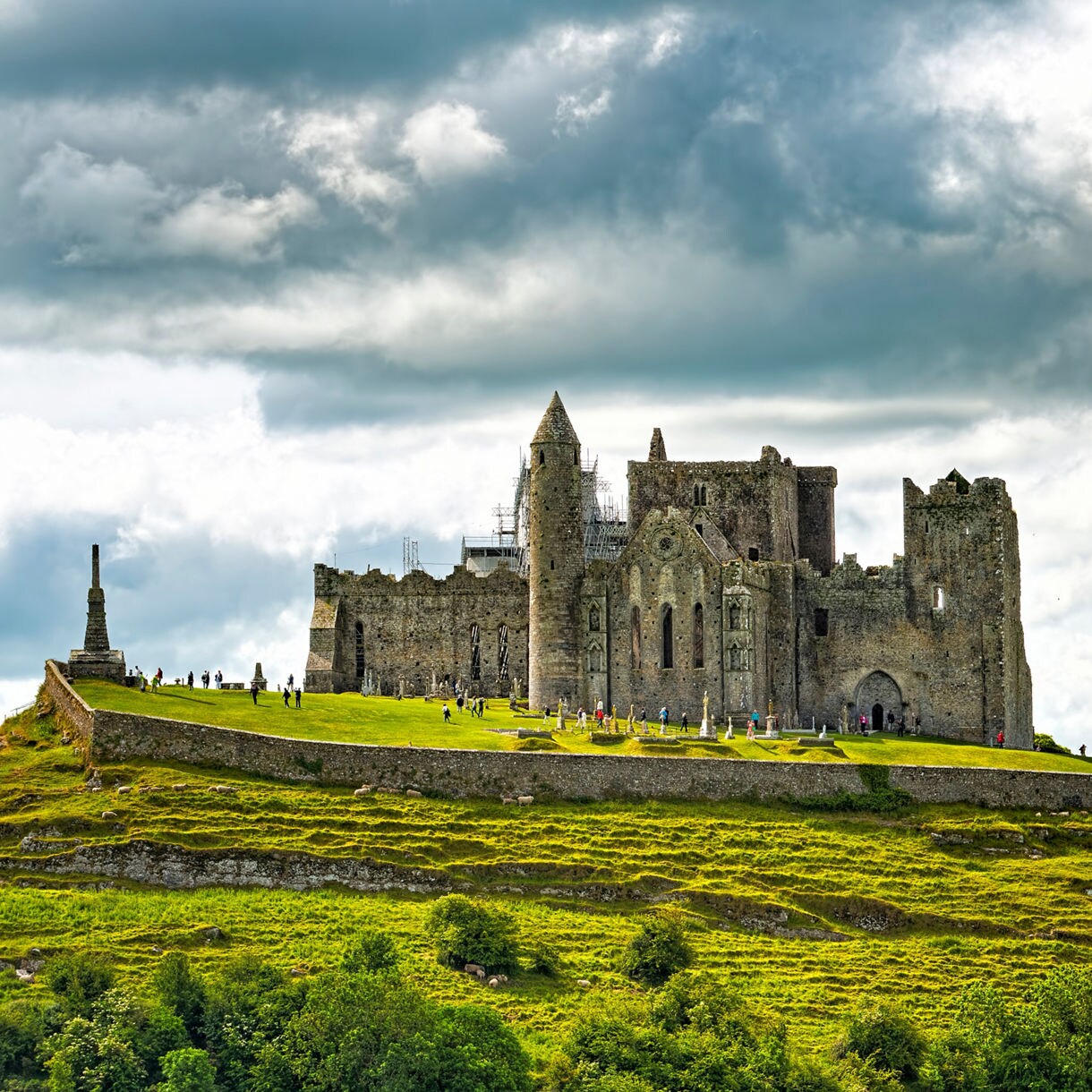 Stone ruins of the Rock of Cashel standing on a grassy hill with visitors scattered across the grounds under dramatic cloudy skies.