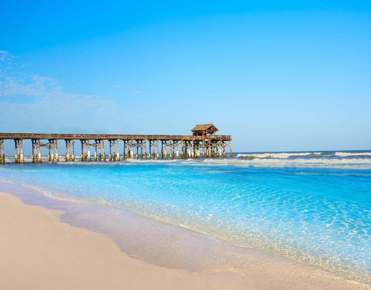 Sandy shoreline at Cocoa Beach with clear blue water and the wooden Cocoa Beach Pier stretching into the ocean under a bright sky.