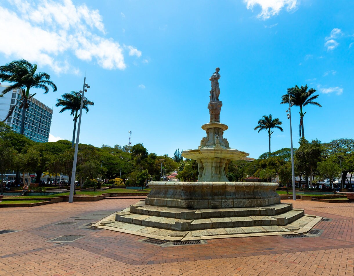 Central stone fountain in Coconut Square, Nouméa, standing on tiered steps with palm trees and a bright blue sky in the background.