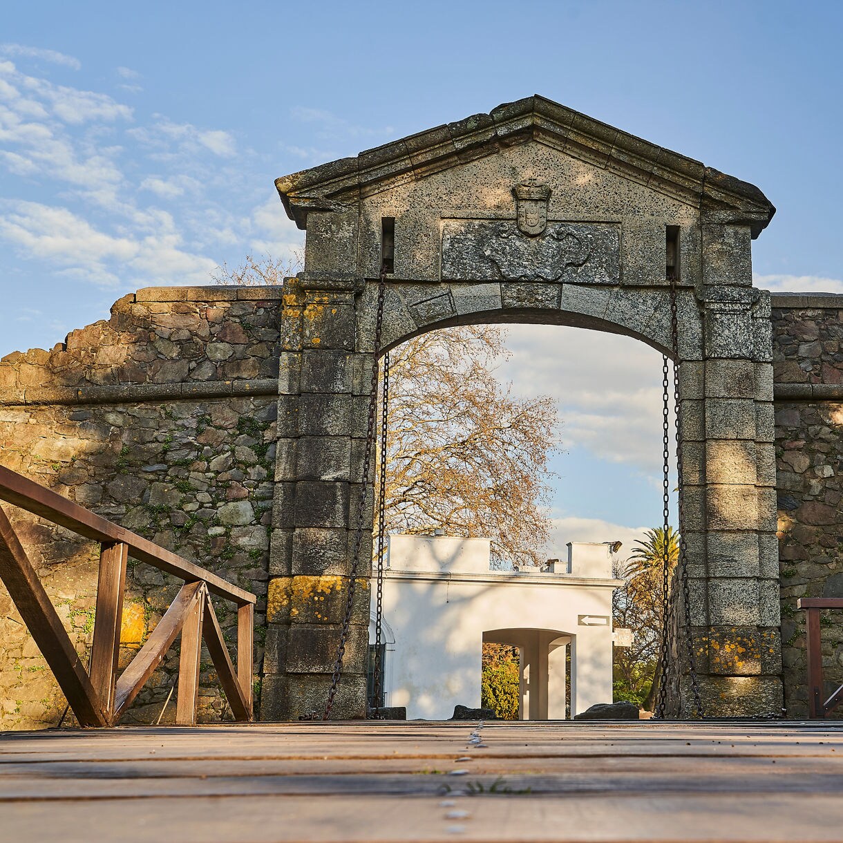 Low-angle view of a stone gate and wooden drawbridge at Colonia del Sacramento, with old fortress walls and a white building framed beyond the arch.
