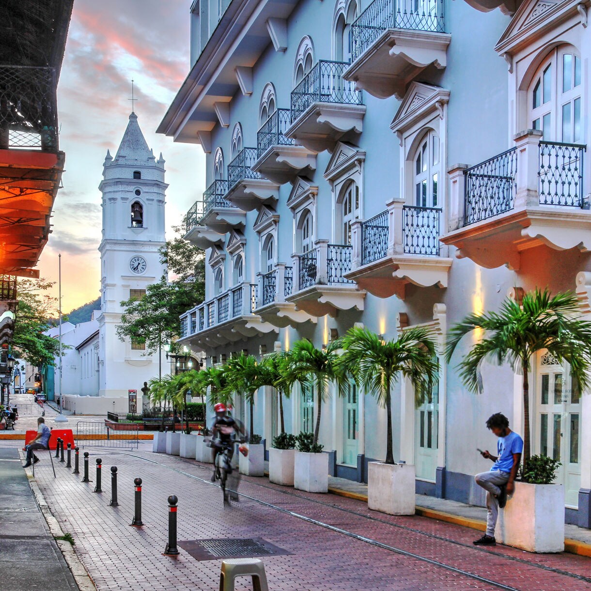 Narrow street lined with pastel colonial buildings, balconies, palm trees and a white church tower in the background at sunset.