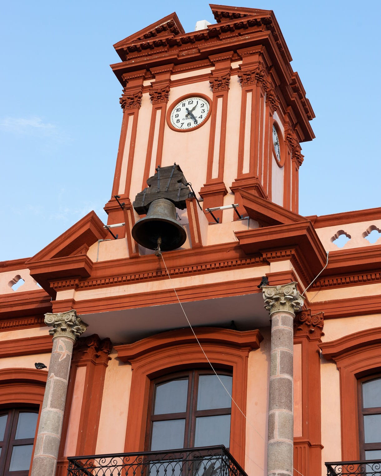 Red and white colonial clock tower with ornate columns and a large bronze bell against a clear blue sky.